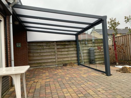 Moderne overkapping met glazen wanden, gepositioneerd op een betegeld terras.